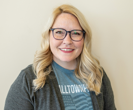 White woman with shoulder length blond hair wearing black rimmed glasses and a light blue shirt t-shirt with white wording and graphic design, and a dark gray cardingan over it.