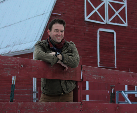 A smiling man in a green jacket and scarf leans on a red wooden fence in front of a large red barn with white trim. There is snow on the ground and the barn roof.