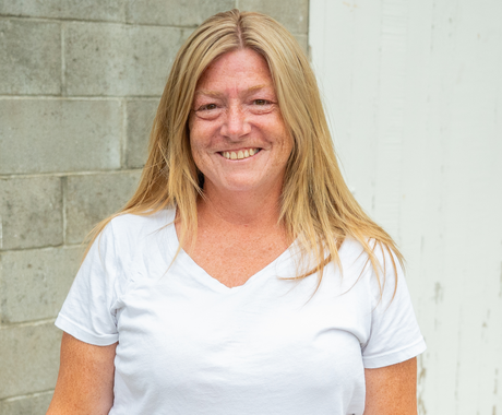 A woman with straight, blonde hair just past her shoulders smiles in front of a grey cinder block wall. She's wearing a white t-shirt.