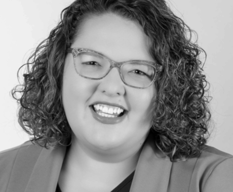 Woman smiling at the camera, with shoulder length curly brown hair, glasses, and a jacket over a dark shirt