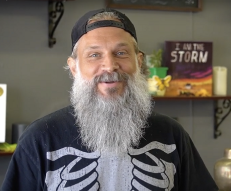 Man with full gray beard and backwards hat in T-shirt with rib cage on the front looking straight at camera