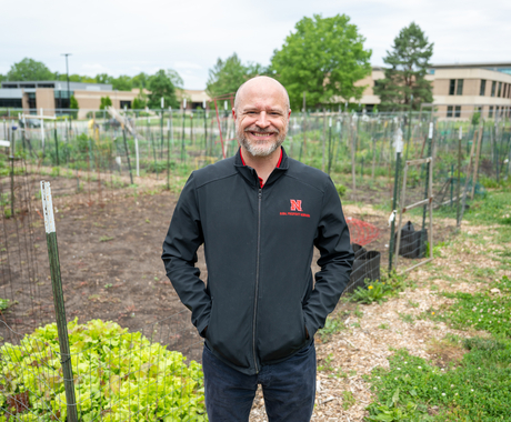 Man with a bald head and facial hair, wearing a zip up collared black shirt with a red "N" on the left breast, smiles at the camera. He is standing in the middle of a garden with leafy greens and sprouts coming up, with fencing around each plot.