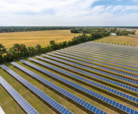 Aerial view of rows of solar panels on top of green grassland bordered with trees on each side. In the background is a farm field, and a farm.