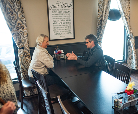 White woman with short blond hair sits across a white man with short dark hair, both sitting at a black long table with black chairs in a room where windows are on each side of them and poster with wording on it on the wall