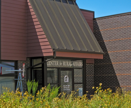 Dark brick building with a glass enclosed entry way. Says "Center for Rural Affairs right under the metal roof overhang, with wildflowers and a windmill in the foreground