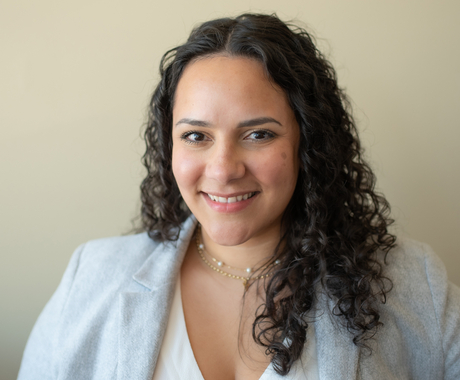 Latina woman with curly black shoulder length hair, wearing a grey business jacket with a white shirt underneath and necklackes around her neck smiles to the camera
