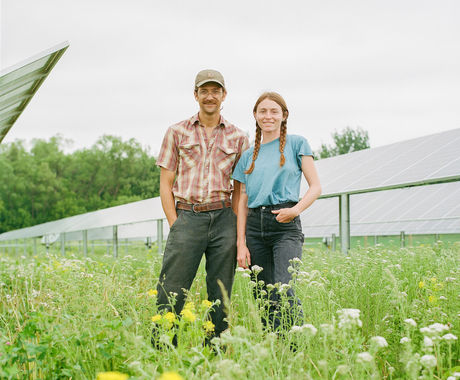 A man and a woman standing in knee-high flowers between rows of solar panels. Man is wearing a short-sleeved red plaid shirt and baseball cap. Woman has red pigtails and is wearing a short-sleeved blue shirt. Both are wearing jeans and smiling directly at the camera.