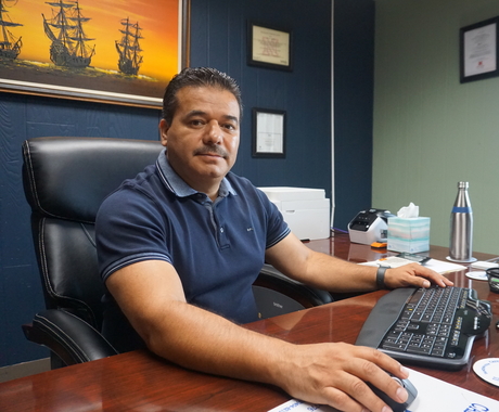 Hispanic male with black hair sits at a desk with a computer and keyboard in front of him, a painting of yellow ships with a yellow background hangs behind him on a wall