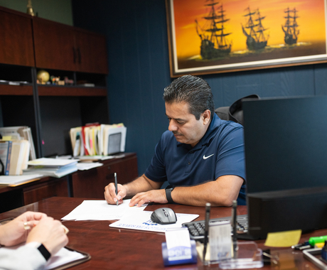 Hombre latino con camisa azul oscura esta sentado detrás de un escritorio con una computadora y firmando un documento en blanco