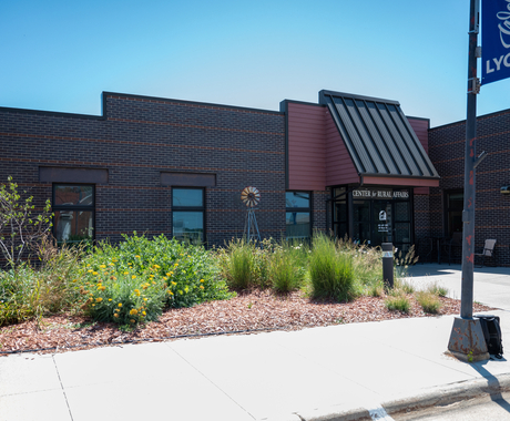Photo of a front of a building with dark red bricks with dark metal sloping to a glass doorway that says "Center for Rural Affairs." In front of the building is a windmill and native grass plantings, with yellow flowers in bloom. In the immediate foreground is a sidewalk and a lamp post with a banner that says "Welcome to Lyons."