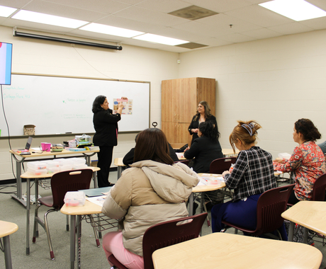 Two hispanic women wearing black outfits stand in front of a classroom of more than 5 women sitting at desks. The woman on the left is holding a paper in her hand and pointing to it.