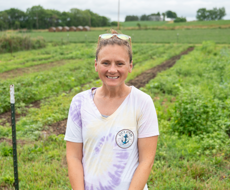 A woman in a white, pink, and light yellow tie-dye t-shirt stands in front of a lush, green field. Her light brown hair is pulled back in a pony tail and she wears sunglasses on her head.