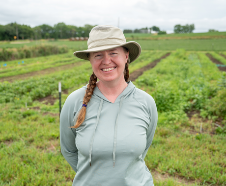 A woman wearing a mint green long sleeved shirt and a beige gardening hat stands in front of a lush green field.