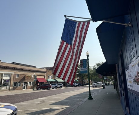 Photo of a flag hanging in the foreground with a downtown main street behind. The main street is brick in the middle, with two story buildings and a handful of cars parked in front