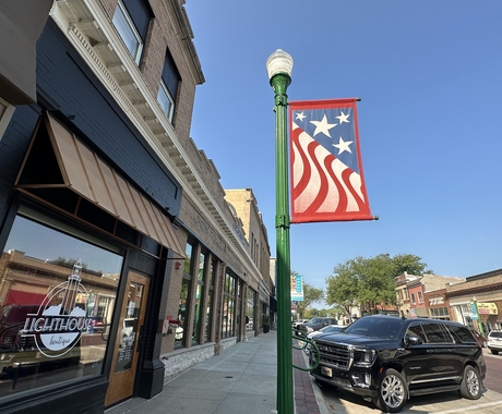 Main street with businesses on the left, a light pole with a flag banner and cars parked along the street