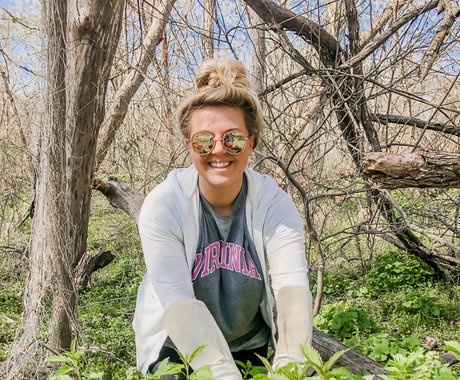 White woman with blond hair wearing sunglasses, white zip up hoodie sweater with a green shirt underneath is kneeled down in greenery
