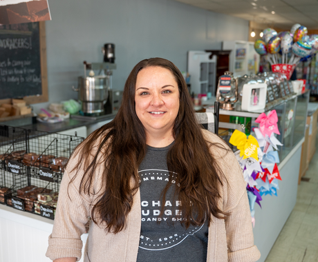 Smiling woman with long dark hair wearing a gray “Homemade Orchard Fudge & Candy Shop” T‑shirt and beige cardigan stands in a bright candy shop interior. Behind her, wire baskets hold fudge, while to her right a rack displays colorful gift bows and festive balloons.
