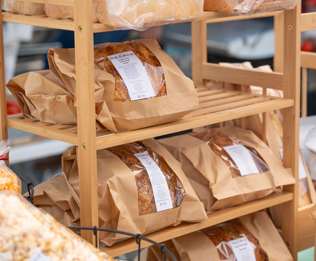 A wooden rack of an assortment of breads, wrapped in clear cellophane and brown parchment. This photo appears to be from a table at a farmers market.