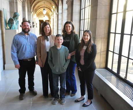 Man in a blue shirt, woman in a brown jacket, boy and woman wearing green shirts and a woman wearing a black jacket stand in the hallway of the Nebraska state capitol.