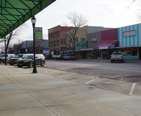 Rural Main Street, with a sidewalk in the foreground, a brick street in the middle, and a row of 2-story buildings with storefronts on the bottom level