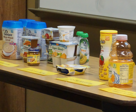 Baby and toddler food sits on a brown table. From left to right, there is Gerber Whole Wheat Cereal,  baby food in plastic containers, Yogurt blends in white plastic cylinders, baby food in a squeezable pouch, Gerbers Graduates puffs in a tall cylinder, and juice.