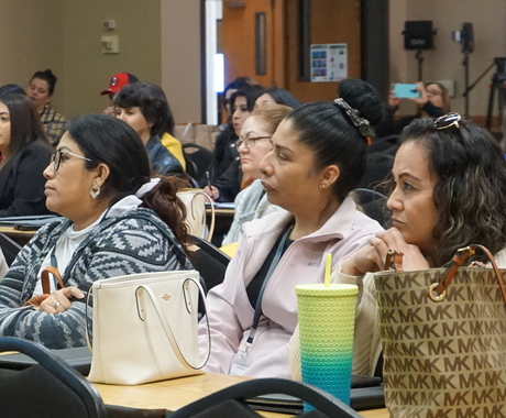 3 Latina women sitting at a table with their purses in front of them, they're surrounded by a room full of people at a conference