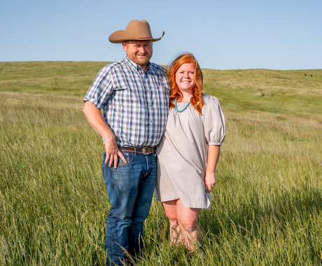A man and woman stand together in a grassy field under a clear blue sky. The man wears a cowboy hat, plaid shirt, jeans, and boots, while the woman wears a light-colored dress with turquoise jewelry and cowboy boots. They are smiling and standing close to each other.