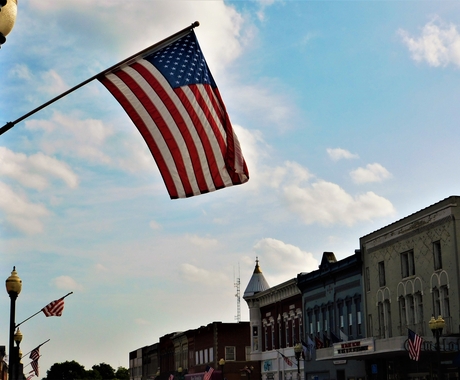 Flag flying from a lamp post on a Main Street. In the background you can see tops of 2-story brick buildings with large windows, and a blue sky with white fluffy clouds