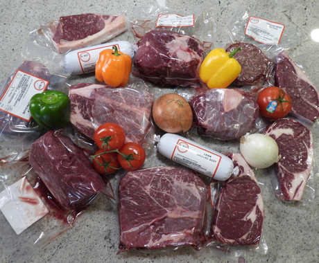 A few cuts of packaged beef arranged in a circle on a table. Cuts are in clear packaging with a couple of white packages of ground beef. Arranged throughout are fresh peppers, tomatoes, and onions.