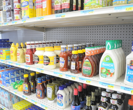 Shelves of food at a small town grocery store. In the foreground is ranch, ketchup, mayo, honey, and to the far left is aluminum foil and baggies.