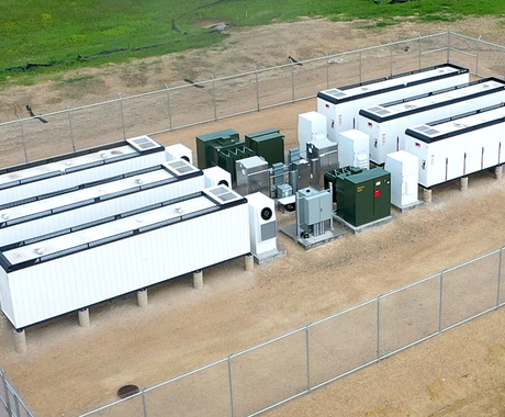 Battery storage project- six small white rectangle buildings (about the size of tractor trailers) lined up three on each side with green and gray electric junction boxes in between. All are in a chain linked fence area with brown dirt underneath.
