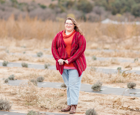 A woman standing in a bare-looking field smiles at the camera. She wears a red cardigan over a red/orange shirt, jeans, and brown boots. In front of her, we see a black tarped row with small lavender plants. We see rows of black tarp with lavender behind her. 