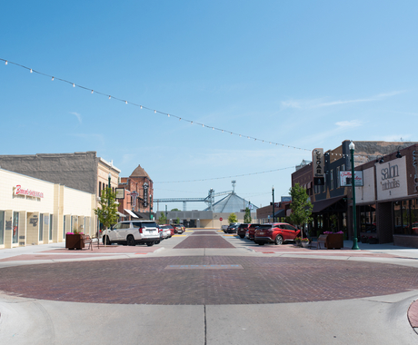 Photo of a small town's main street, with a large brick circle in the middle of an intersection, then brick one-story buildings on either side, with a grain bin in the background, at the end of the main street road.