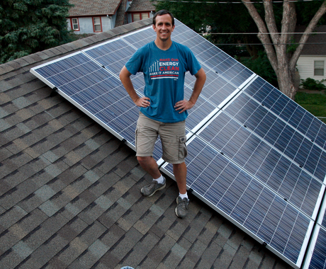 A white man with dark hair stands on a grey shingled roof, in front of newly installed solar panels. He is smiling with his hands on his hips, wearing a light blue T-shirt that says "Make our energy clean, make it American" and khaki shorts.