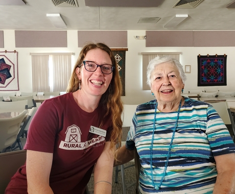 Two women, the right is in  a maroon shirt, long red hair, and glasses. The one on the left is older, wearing a striped blue and white shirt with short white hair.