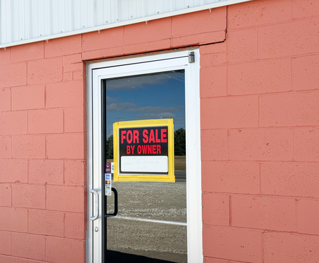 Salmon-colored brick building with a clear business door in middle. A "for sale by owner" sign in the top center of the door.