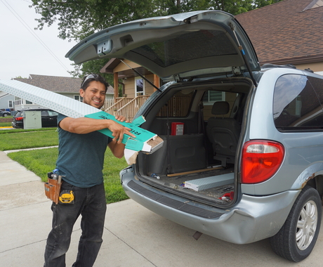 Hispanic man wearing a navy shirt and black jeans carries box of white box of wood flooring panels out of the back of a light blue van outside a neighborhood
