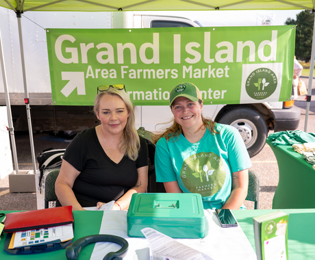 Two women sit under a green canopy at a table covered with a green tablecloth. The woman on the left has long, blonde hair and wears a black v-neck t-shirt and sunglasses on her head. The woman on the right wears a teal t-shirt that reads "GRAND ISLAND FARMERS MARKET" and a green baseball cap. The sign behind them says "GRAND ISLAND Area Farmers Market Information Center."