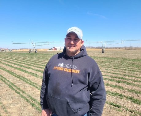 Un hombre con una sudadera negra con capucha que dice "Elite Seed Treatment" y una gorra blanca de "Center for Rural Affairs" está de pie en medio de un campo. En el suelo se ven hileras verdes de plantas con rastrojo entre ellas. Al fondo se aprecia un sistema de riego por pivote, un gran cobertizo y cielo azul.