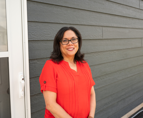 Woman in a short sleeved, red/orange blouse standing in front of a white door and gray siding. She is smiling warmly and looking into the camera.