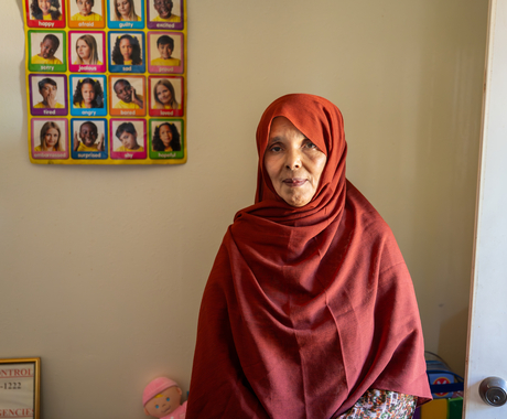 A dark-skinned older woman wearing a maroon hijab, looking directly at the camera. She is standing in front of a beige wall with a white door to her left (right side of the photo). On the left side is a poster hanging up that says "Emotions" and has photos of children and words that state emotions in each photo.