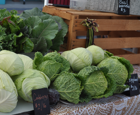 A display of fresh vegetables at a farmers market stand, including heads of green cabbage and savoy cabbage arranged on a patterned tablecloth. Small chalkboard signs indicate prices: "Cabbage $2/lb," "Eggplant $3/lb," and "Basil (varieties) $3." Other leafy greens and produce are visible in the background, along with wooden crates and metal containers.
