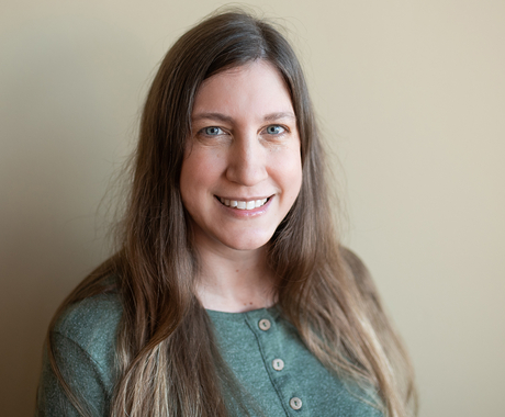 White woman with long brown hair wearing a light green shirt with three buttons down the middle stands in front of a beige wall smiling and posing for the camera.