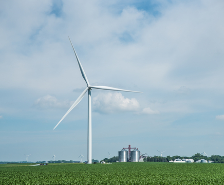 Turbina eólica en un campo agrícola con cultivos a su alrededor y operaciones agrícolas al fondo.