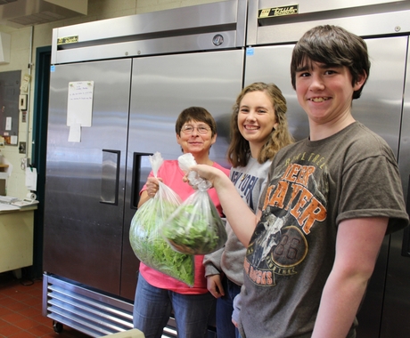 A lunch lady in a pink shirt holds a big bag of freshly cut lettuce, accompanied by two high school students - a boy and a girl - standing in front of industrial sized refrigerators in a school kitchen