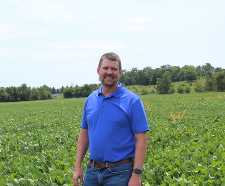 Man with short hair and facial hair smiles at the camera. He is standing in the middle of a soybean field. The beans are leafy green with purple flowers. The man is wearing a collared short sleeve blue shirt.