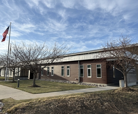 Photo of a one story brown building with tall windows. In the foreground is short green grass. Two flags fly in front of the main door. Above the building is blue sky with white wispy clouds.