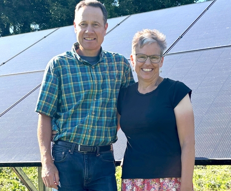 Man and woman stand with their arms around each other, in front of a solar array. They stand in bright green grass with green leafy trees behind.