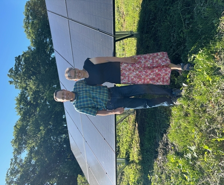 Man and woman stand with their arms around each other, in front of a solar array. They stand in bright green grass with green leafy trees behind.