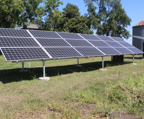 Un panel solar, con césped verde debajo y una hilera de pinos más un contenedor de granos detrás.